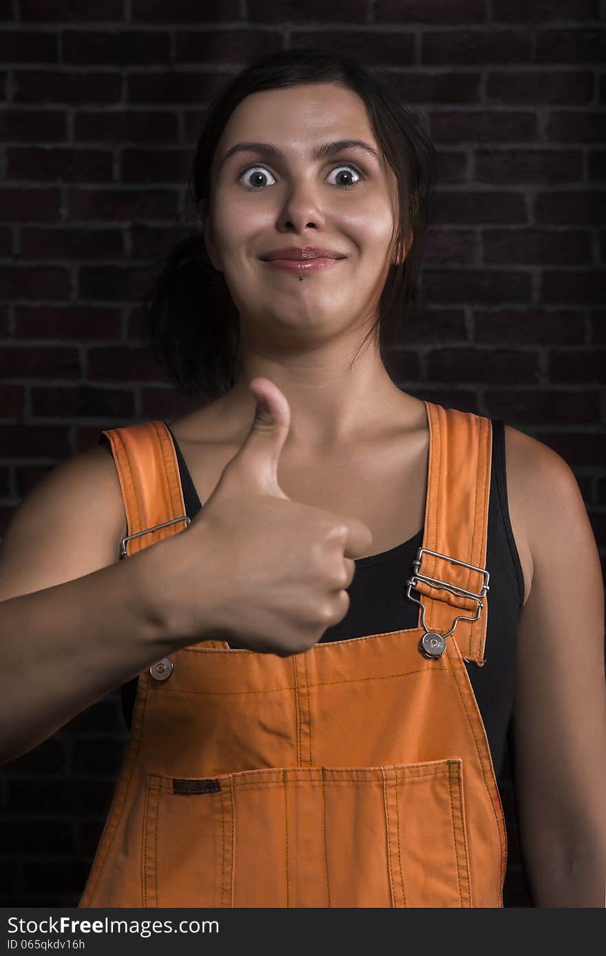 Portrait of young pretty brunette girl, wearing bright orange color overalls, in great mood showing thumbs up gesture, with big smile and wide open eyes on her face. Brick wall background. Portrait of young pretty brunette girl, wearing bright orange color overalls, in great mood showing thumbs up gesture, with big smile and wide open eyes on her face. Brick wall background.