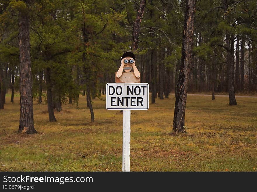 A "Do Not Enter" sign with a man with binoculars carved on top of the sign surrounded by pine trees. A "Do Not Enter" sign with a man with binoculars carved on top of the sign surrounded by pine trees