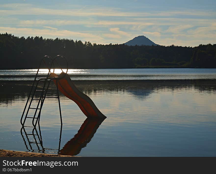 Abandoned old children slide stays in pond at the end of summer , reflection in blue water level, forest at horizon.