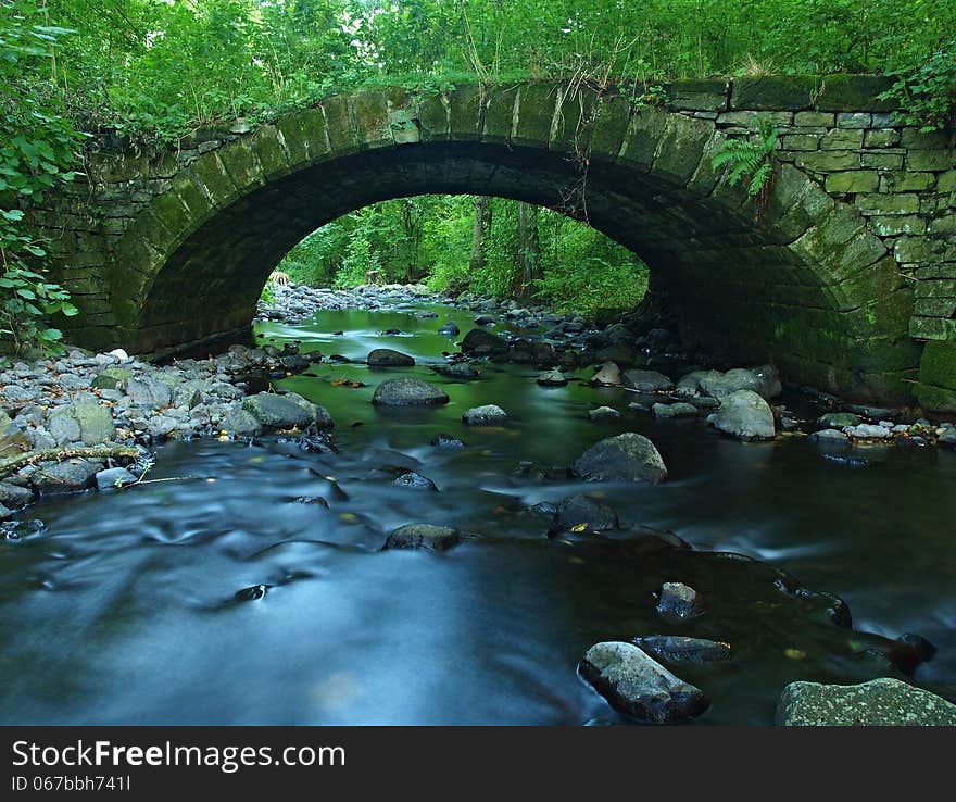 The Old Stony Bridge Of Mountain Stream In Leaves Forest, Blue Blurred Water Is Below