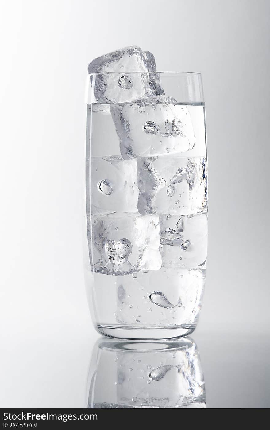 Glass filled with water and ice cubes on a white/grey background, reflection at the bottom. Glass filled with water and ice cubes on a white/grey background, reflection at the bottom