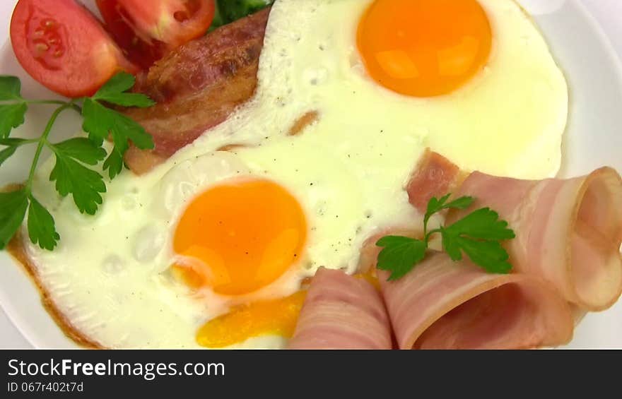 White plate with two fried eggs, bacon, sliced tomato and cucumber, parsley leaves rotates on a white background. Close-up. White plate with two fried eggs, bacon, sliced tomato and cucumber, parsley leaves rotates on a white background. Close-up