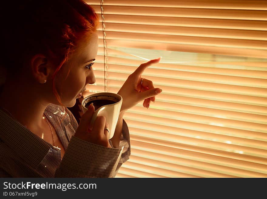 Woman with coffee stares through blinds early morning