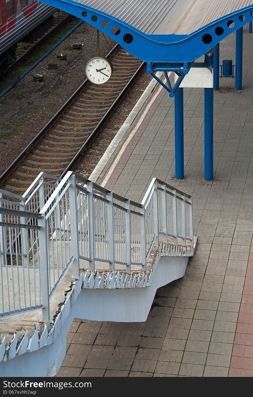 Railway platform and clock at the railway station without passengers and trains. Urban landscape - perspective way on a railroad with stairway.