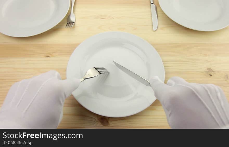 Wooden table. White plate. Hands in white gloves with a fork and a knife cut a piece of invisible food. Wooden table. White plate. Hands in white gloves with a fork and a knife cut a piece of invisible food