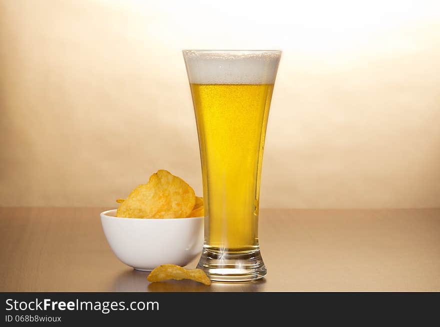 Glass of light amber beer and bowl with chips against brown wall