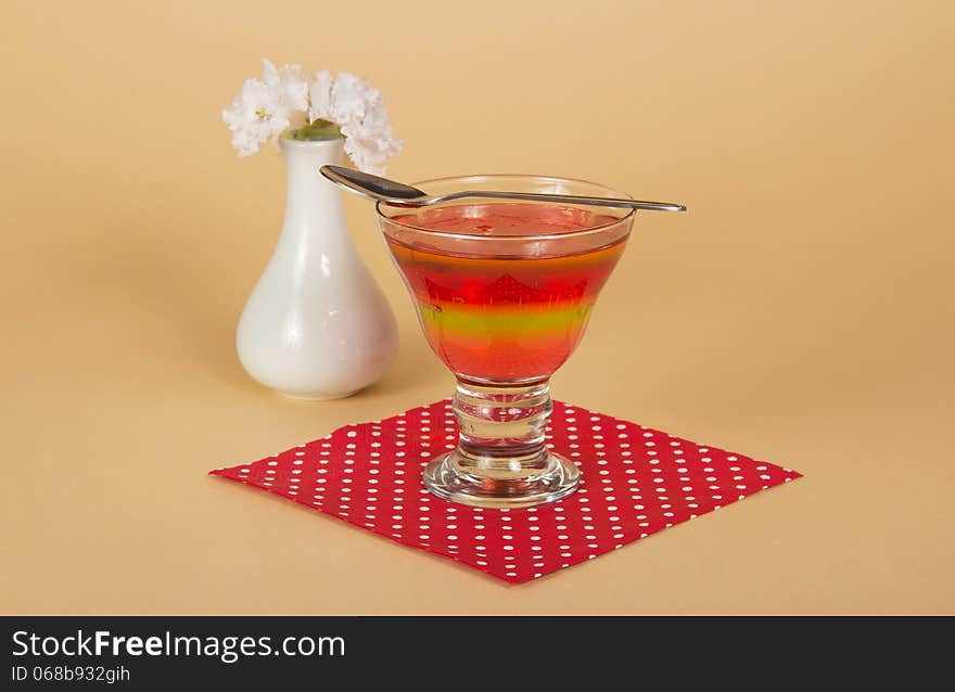 Glass with jelly and a spoon on a napkin, a vase with the flowers, on a beige background. Glass with jelly and a spoon on a napkin, a vase with the flowers, on a beige background