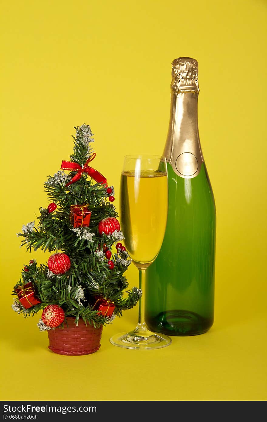 Bottle and wine glass with champagne, and a small fir-tree in a pot on a yellow background