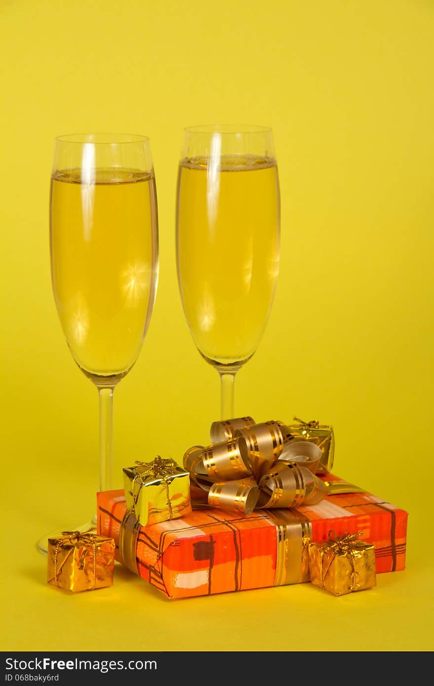 Wine glasses with champagne, a gift box with a beautiful bow and souvenirs on a yellow background. Wine glasses with champagne, a gift box with a beautiful bow and souvenirs on a yellow background