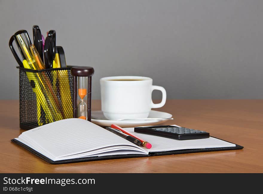 Open notepad, office accessories in a support the calculator, hourglasses and a cup of coffee on a table. Open notepad, office accessories in a support the calculator, hourglasses and a cup of coffee on a table