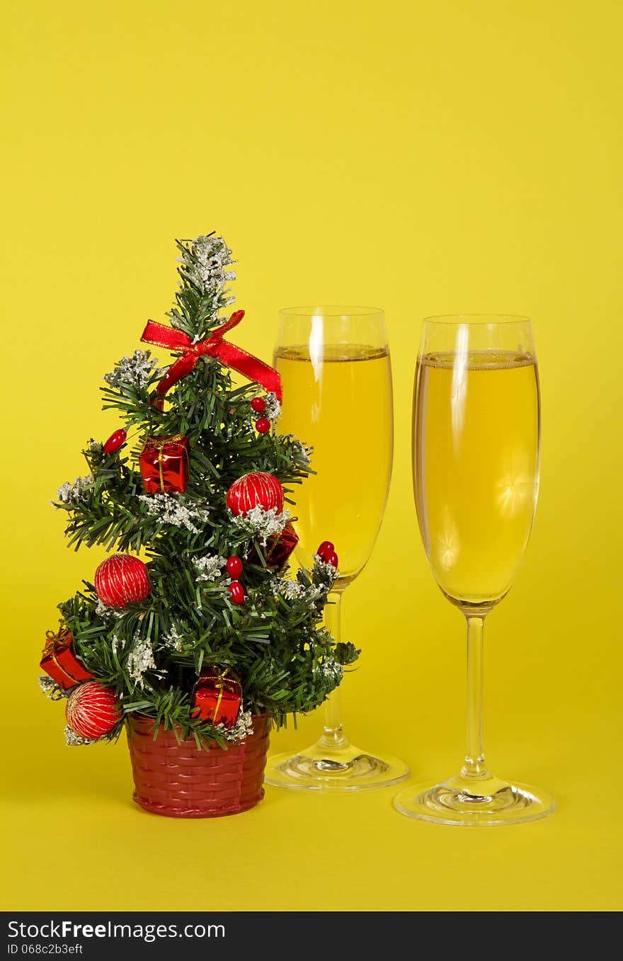 Small Christmas fir-tree in a pot, and two wine glasses with champagne on a yellow background