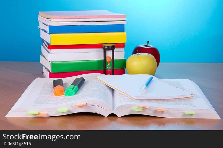 The open book with bookmarks, exercise book a pen and markers, a pile of the closed books a hourglass and apples on a table