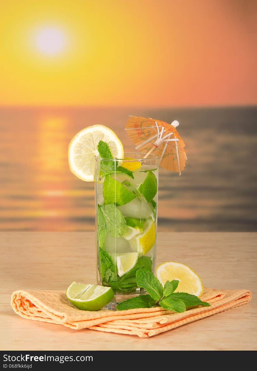 Mojito with umbrella, a lemon, napkin on a table against the sunset. Mojito with umbrella, a lemon, napkin on a table against the sunset