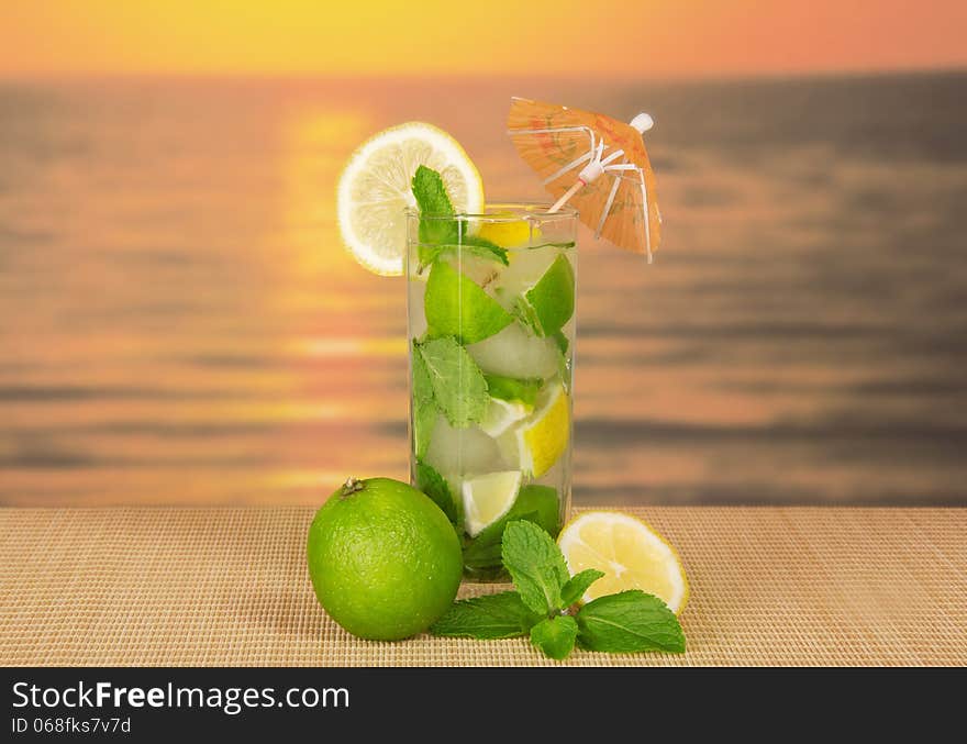 The glass of a cold mojito, is decorated with an umbrella, the spearmint, on a bamboo cloth against the sunset. The glass of a cold mojito, is decorated with an umbrella, the spearmint, on a bamboo cloth against the sunset