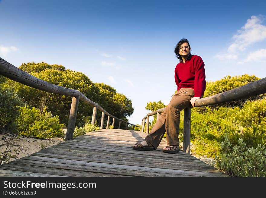 Man sitting on the rustic wooden railings of a coastal boardwalk crossing the dunes and beach sand, as he relaxes and enjoys the sunshine and the tranquillity of nature. Man sitting on the rustic wooden railings of a coastal boardwalk crossing the dunes and beach sand, as he relaxes and enjoys the sunshine and the tranquillity of nature.