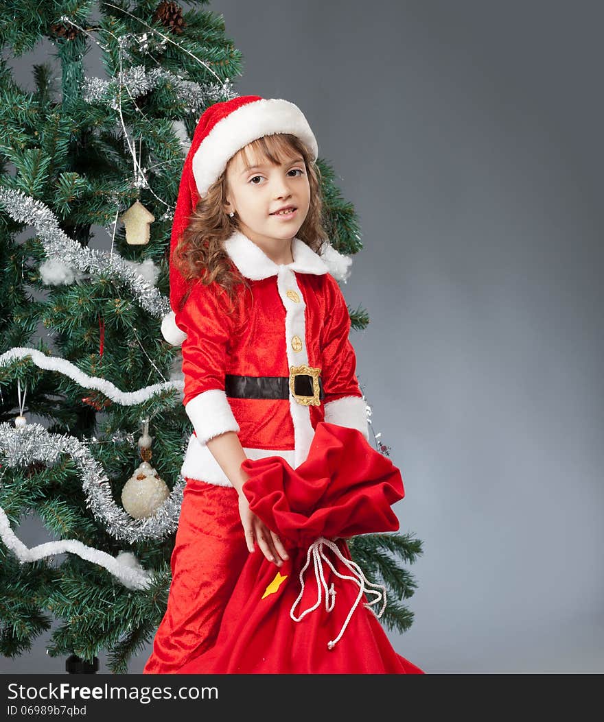 A little girl dressed in a suit Snta Claus standing near a Christmas tree and holding a red bag with gifts. A little girl dressed in a suit Snta Claus standing near a Christmas tree and holding a red bag with gifts