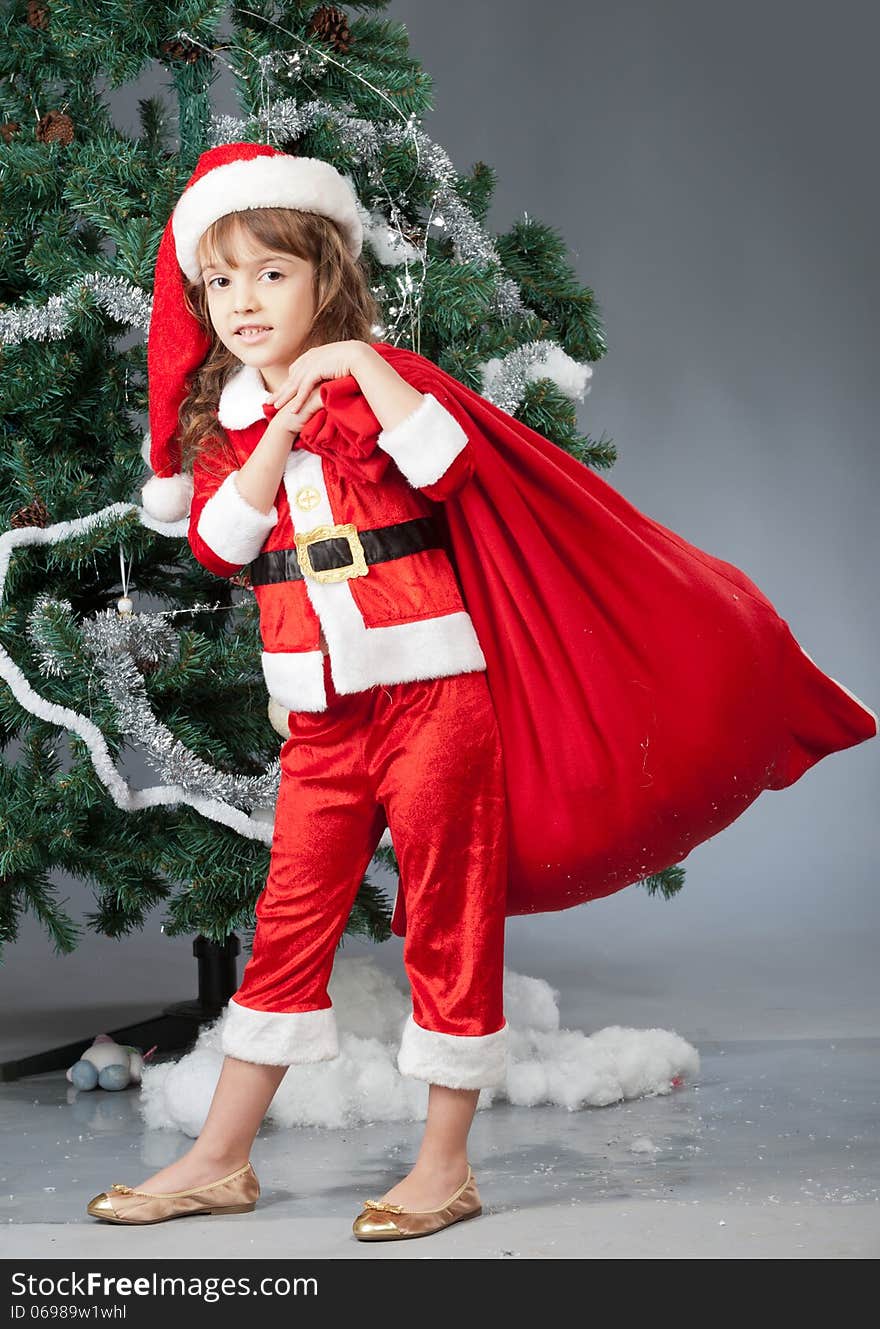 A little girl dressed in a suit Snta Claus standing near a Christmas tree and holding a red bag with gifts. A little girl dressed in a suit Snta Claus standing near a Christmas tree and holding a red bag with gifts