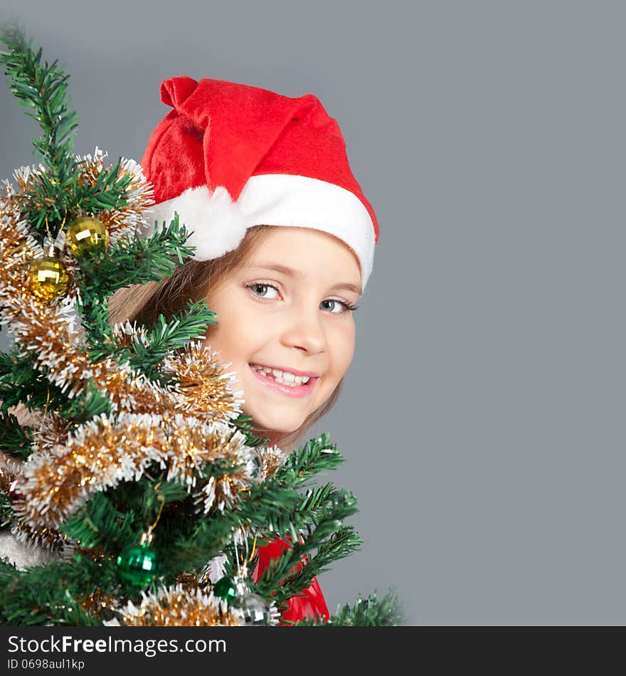 Little girl in Santa hat smiling and peeking out from behind the Christmas tree. Little girl in Santa hat smiling and peeking out from behind the Christmas tree