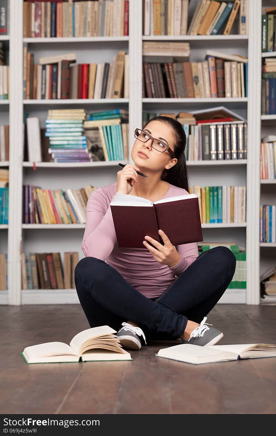 Thoughtful young woman holding a book and looking away while sitting on the floor at the library. Thoughtful young woman holding a book and looking away while sitting on the floor at the library