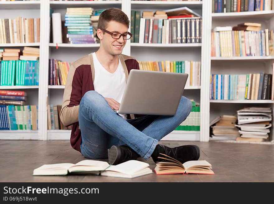 Cheerful young man typing something on laptop while sitting on the floor at the library. Cheerful young man typing something on laptop while sitting on the floor at the library