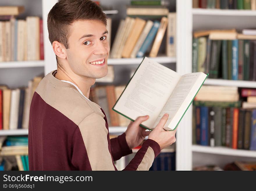 Cheerful young man holding a book and looking over shoulder while standing at the library. Cheerful young man holding a book and looking over shoulder while standing at the library