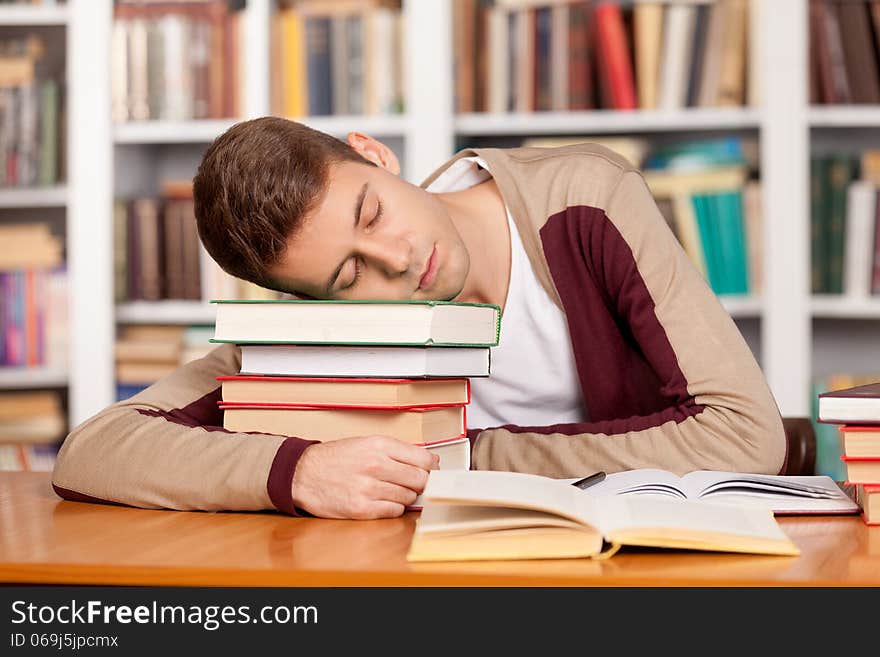 Tired young man holding his head on the book stack and sleeping while sitting at the library desk. Tired young man holding his head on the book stack and sleeping while sitting at the library desk