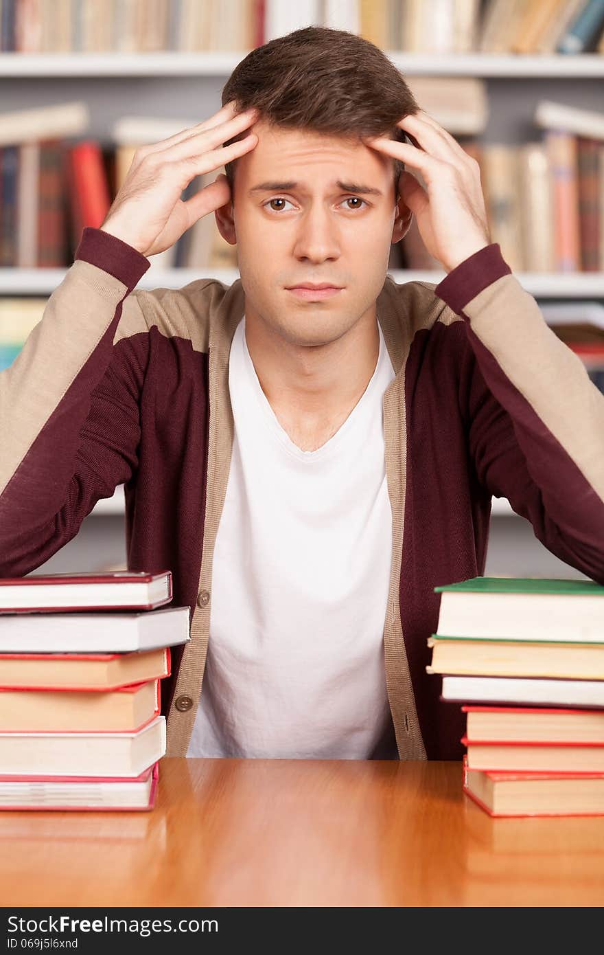 Tired young man holding his head in hands and leaning at the book stack while sitting at the library desk. Tired young man holding his head in hands and leaning at the book stack while sitting at the library desk