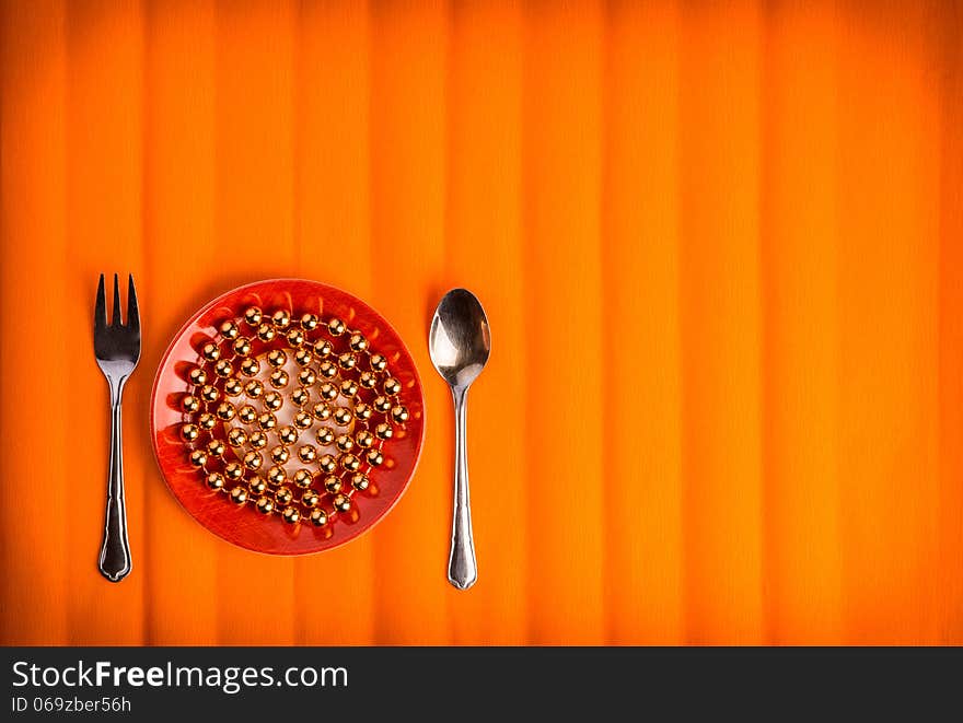 One lovely plate with golden beads on it , little fork and spoon by sides. Over textured bright orange color background. One lovely plate with golden beads on it , little fork and spoon by sides. Over textured bright orange color background.