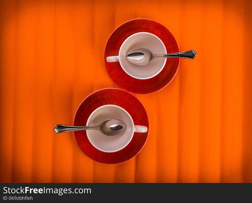 Two white empty cups with tea spoons, on red plates over orange color background, view from above