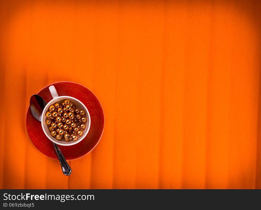 One lovely cup with golden beads in it , little fork and spoon by sides. Over textured bright orange color background. One lovely cup with golden beads in it , little fork and spoon by sides. Over textured bright orange color background.