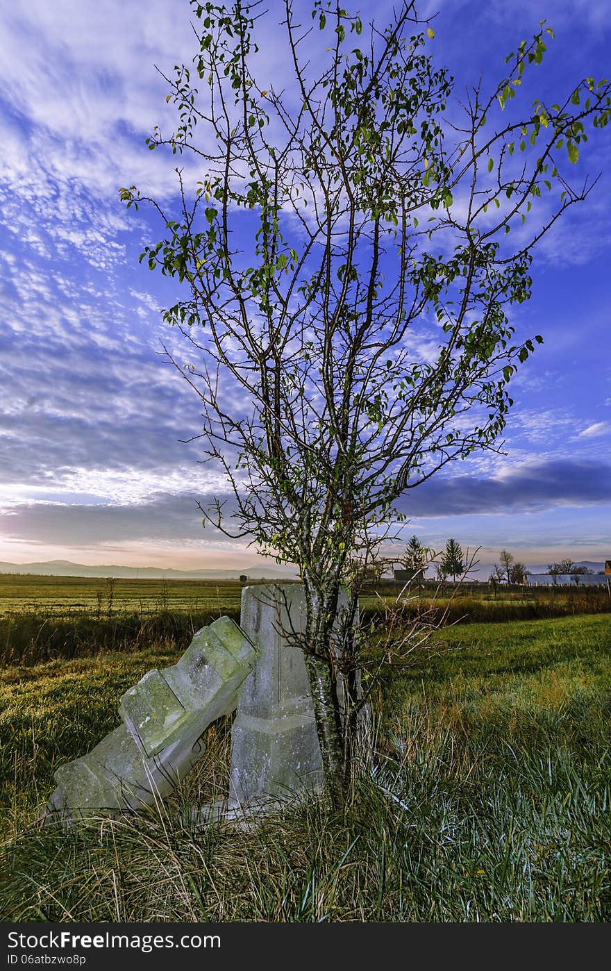 Tree and tomb wtih a cross near the road - for someone who died in car accident - red dawns background and beautiful blue sky landscape. It is very odd because this tree has grown adjacent to the cross. Tree and tomb wtih a cross near the road - for someone who died in car accident - red dawns background and beautiful blue sky landscape. It is very odd because this tree has grown adjacent to the cross.