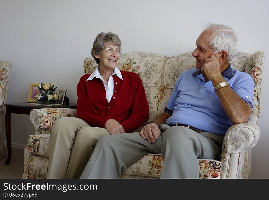 An elderly couple sit and relax on a couch as engage in conversation and enjoy each others company.