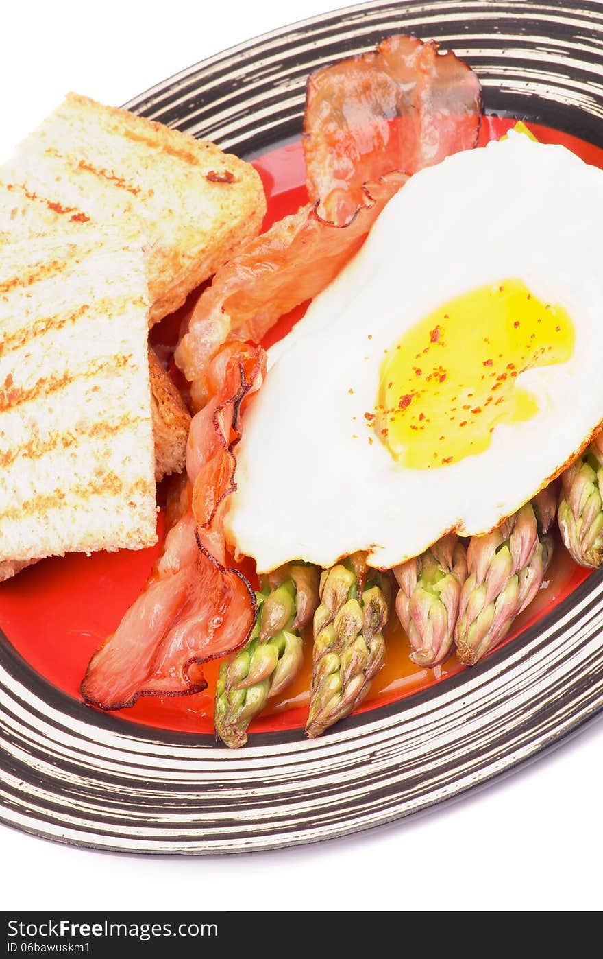 Tasty Breakfast with Fried Egg Sunny Side Up on Delicious Asparagus Sprouts, Roasted Bacon and Toasts on Red Striped Plate closeup on white background