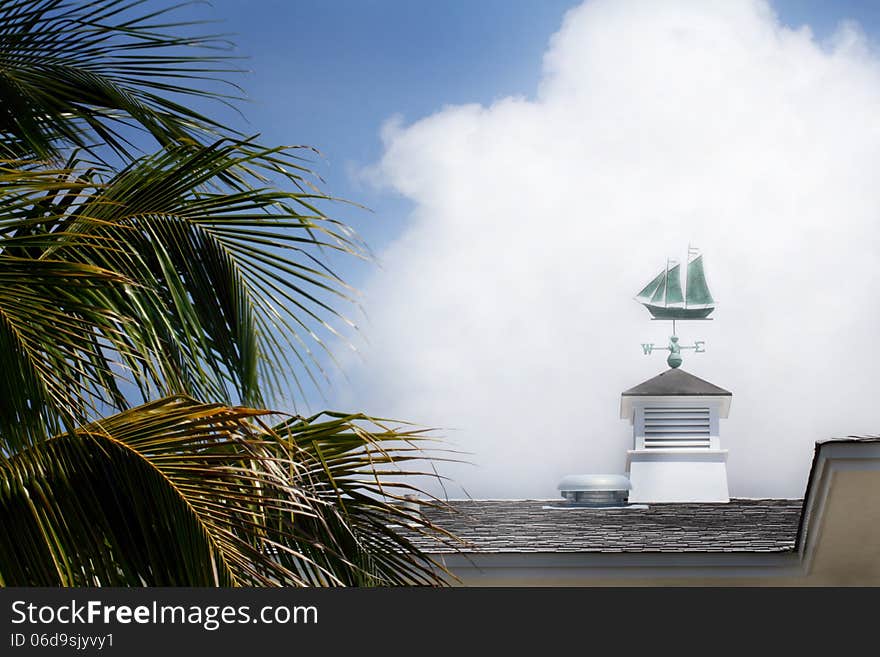 A green brass sailboat nautical weather vane on a rooftop cupola with fluffy clouds in the background and some palm branches in the foreground. Blue sky. A green brass sailboat nautical weather vane on a rooftop cupola with fluffy clouds in the background and some palm branches in the foreground. Blue sky.