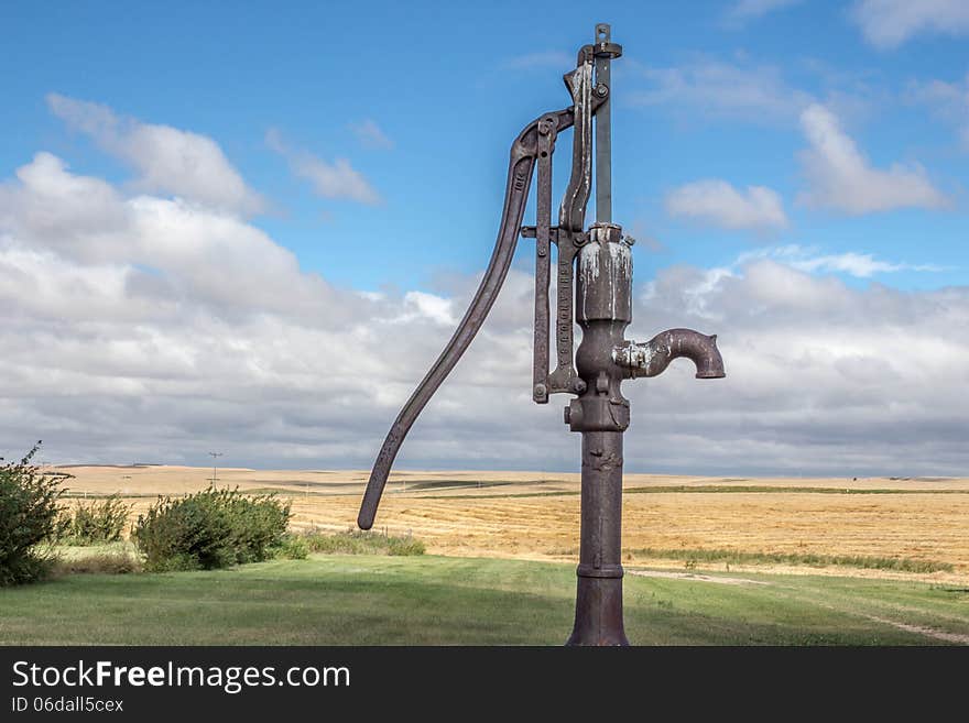 Old rusty antique water pump sitting in a field of golden wheat and green grass and a bright blue cloud filled sky. Old rusty antique water pump sitting in a field of golden wheat and green grass and a bright blue cloud filled sky