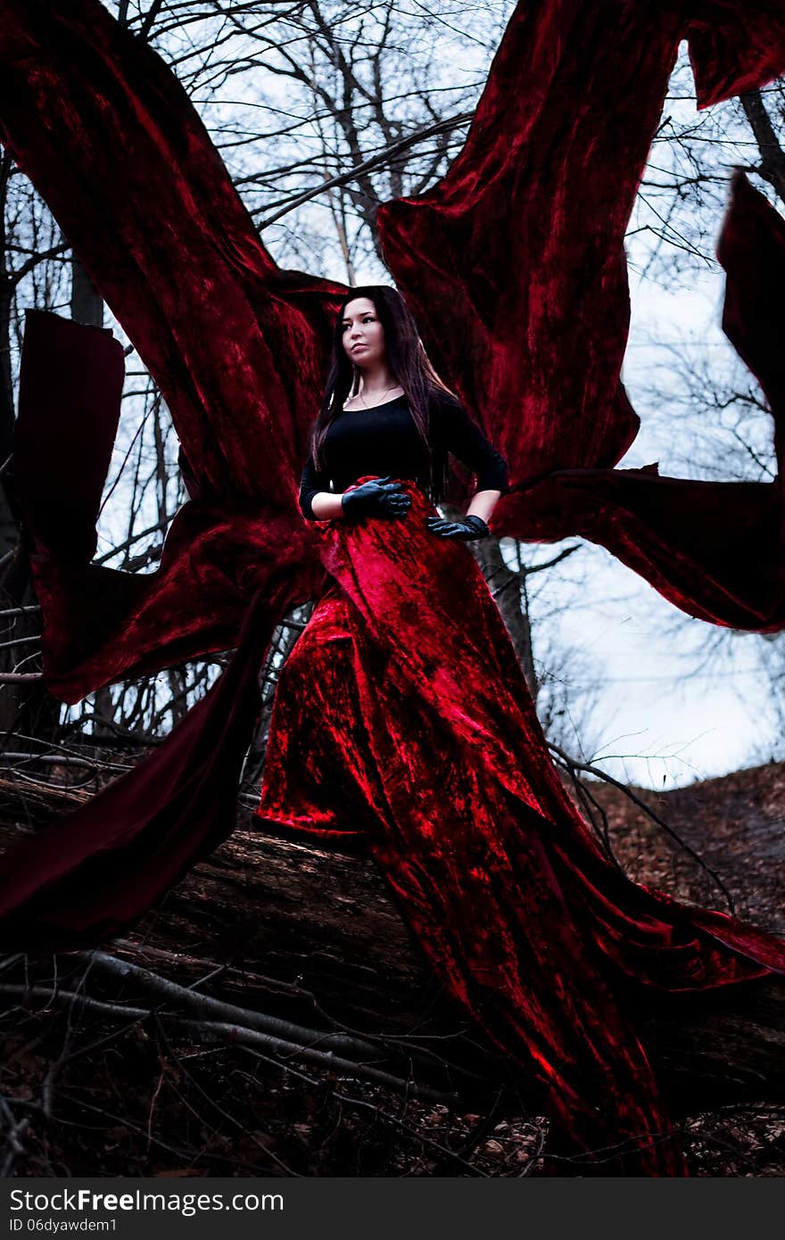 Mysterious woman or witch in long red dress standing in dark forest with flying fabric