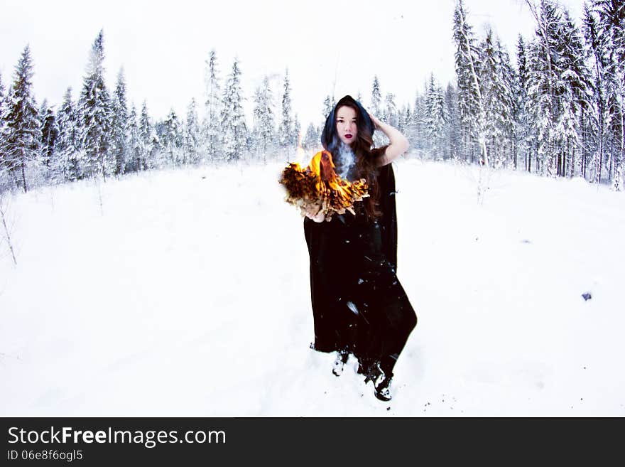 Witch or woman in black cloak with fire ball in white snow forest