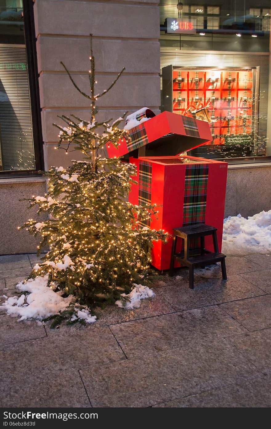 Small christmas tree snow covered in a small square of Zurich with a gift box behind