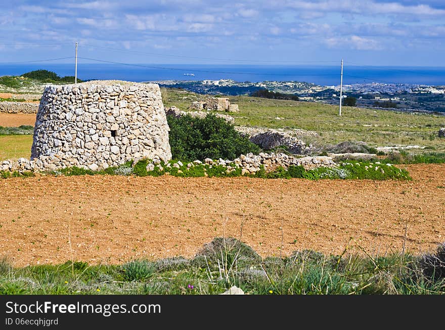 A corbel led stone hut and a cultivated field with crops in the maltese countryside. In the background is the island of Gozo visible surrounded by clear blue Mediterranean sea water. A corbel led stone hut and a cultivated field with crops in the maltese countryside. In the background is the island of Gozo visible surrounded by clear blue Mediterranean sea water.