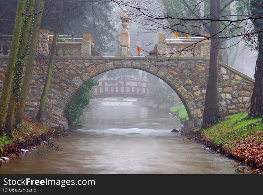 The photograph shows a small river flowing through the park. Passed over the river is a stone bridge for pedestrians. The photograph shows a small river flowing through the park. Passed over the river is a stone bridge for pedestrians.