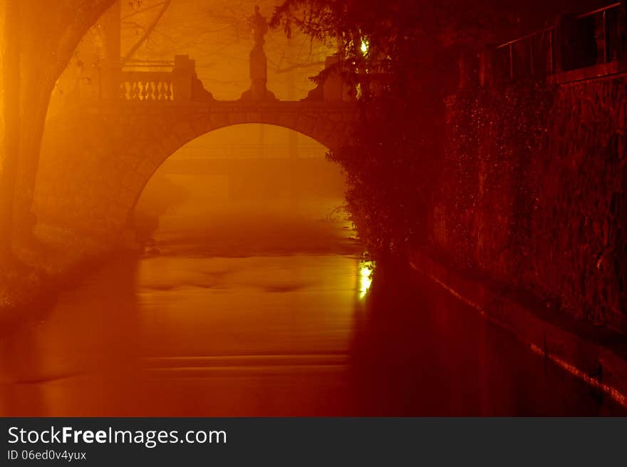 The photograph shows a small river over which passed the arched stone bridge. It is a misty night. Glow illuminates the dark area on the left side of the cadres of electric lamps. The photograph shows a small river over which passed the arched stone bridge. It is a misty night. Glow illuminates the dark area on the left side of the cadres of electric lamps.