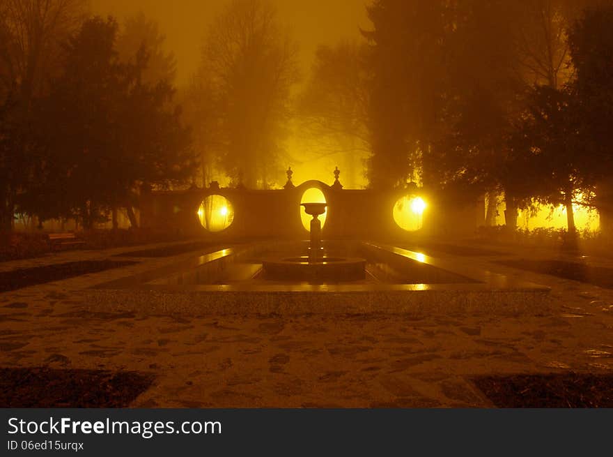 The photograph shows the park at night. Light illuminates the darkness of the park lamps. Above ground fog rises. In the scene we see the brick, corrugated wall with oval holes.