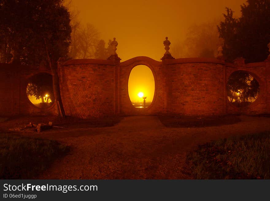 The photograph shows the park at night. Light illuminates the darkness of the park lamps. Above ground fog rises. In the scene we see the brick, corrugated wall with oval holes.