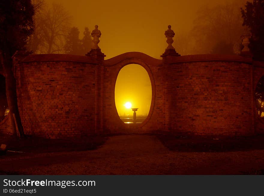 The photograph shows the park at night. Light illuminates the darkness of the park lamps. Above ground fog rises. In the scene we see the brick, corrugated wall with oval holes.