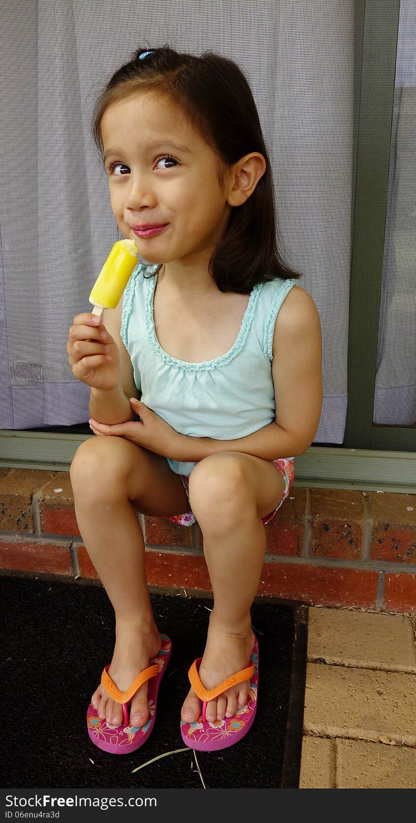 A young Asian girl sits on an outdoor step and grins as she takes a bite of her very cold icy-pole. A young Asian girl sits on an outdoor step and grins as she takes a bite of her very cold icy-pole.