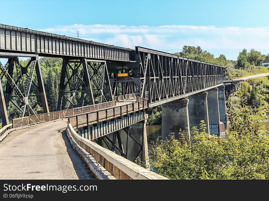 Black steel car bridge stretching across the river,surrounded by trees on a bright summer day covered by blue sky. Black steel car bridge stretching across the river,surrounded by trees on a bright summer day covered by blue sky