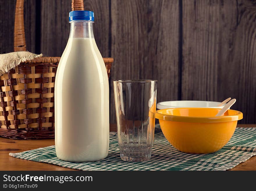 Still life of plastic bottle of milk, empty glass, two plastic bowls and wicker basket over checked table-napkin. Wooden wall background. Still life of plastic bottle of milk, empty glass, two plastic bowls and wicker basket over checked table-napkin. Wooden wall background.