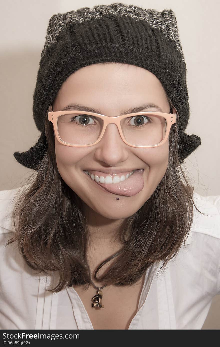 Close-up portrait of young cheerful girl showing her tongue. She is wearing funny black color winter hat, pinky glasses and white shirt. Isolated. Close-up portrait of young cheerful girl showing her tongue. She is wearing funny black color winter hat, pinky glasses and white shirt. Isolated