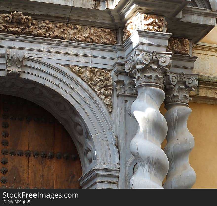 Andalusian architectural doorway detail, showing intricately carved pillars of smooth stone, topped with gold and stone lintels, surrounding heavy, wooden, studded doors