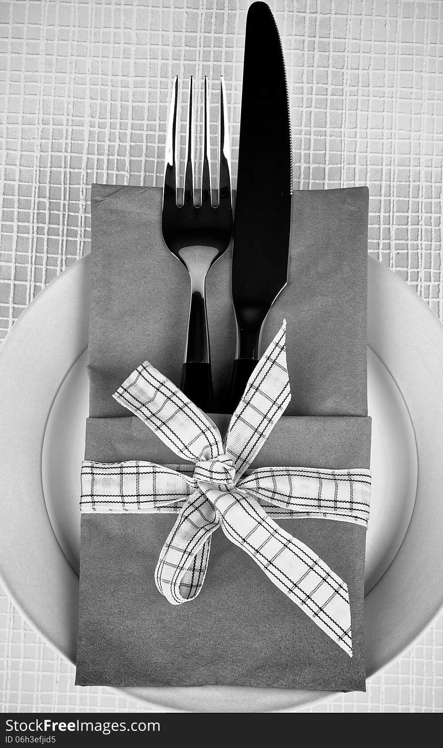 Elegant Table Setting with Fork and Table Knife into Napkin Decorated with Checkered Bow on Plate. Monochrome View